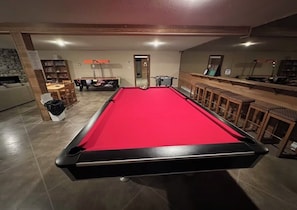 Una mesa de billar con superficie de fieltro rojo, una zona de bar con taburetes y un comedor con sillas y una mesa.