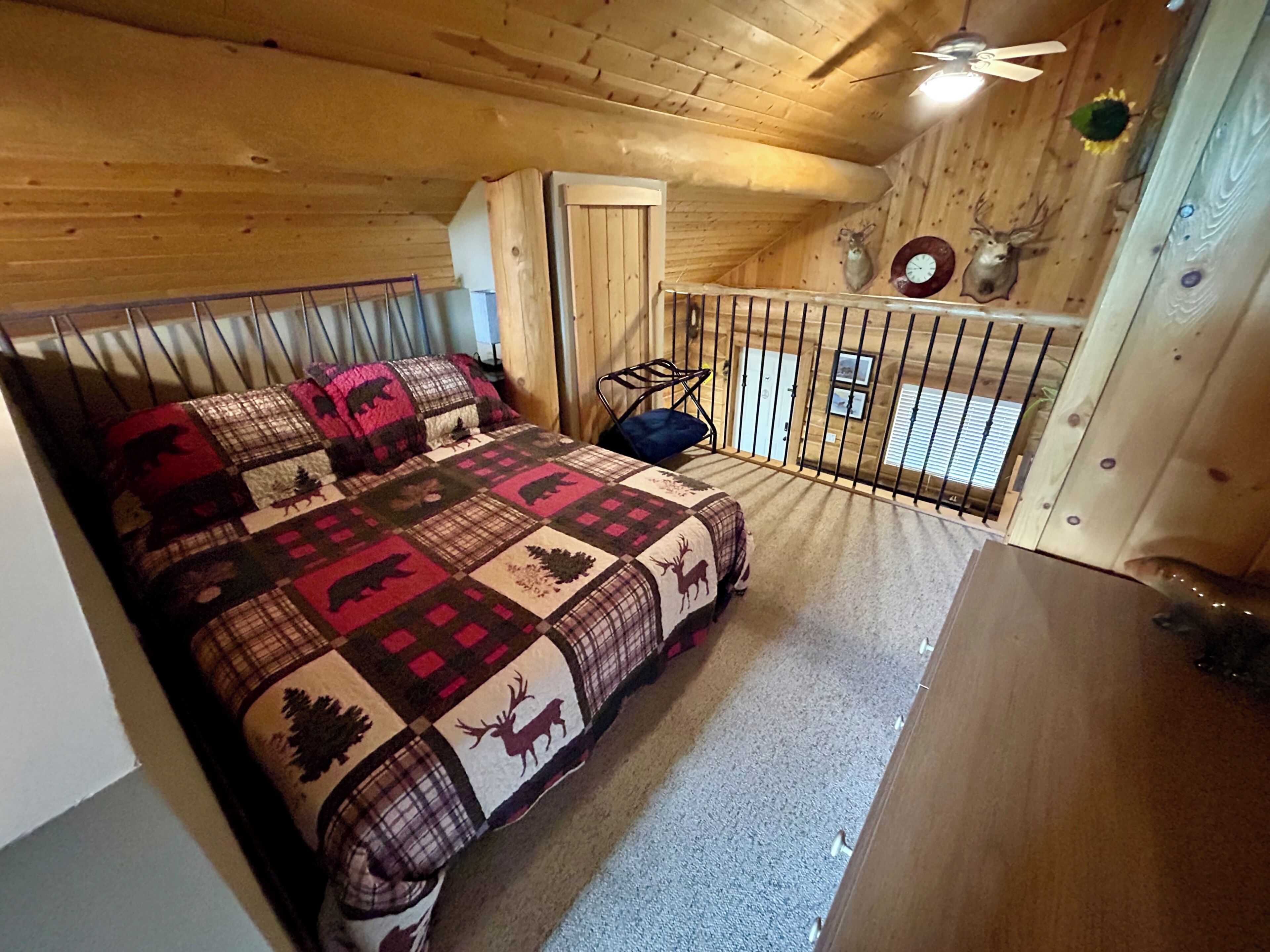 A cozy room with a bed, a chair, a small table, a balcony with a railing, and a ceiling fan.