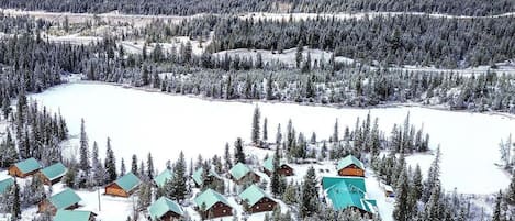 A snow-covered landscape with cabins clustered around a lake, surrounded by dense forests.