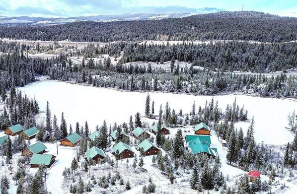 Un paysage enneigé, avec des chalets regroupés autour d’un lac, le tout entouré de forêts denses.