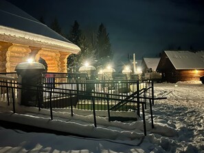 A snow-covered outdoor area with a wooden cabin and a stone structure with lights.
