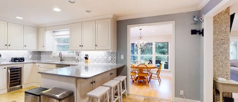A modern kitchen with white cabinets, a large island with stools, a built-in oven, and a dining area with a chandelier.