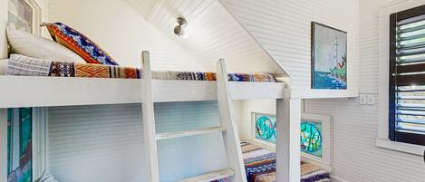 A bunk bed room with a ladder, patterned bedding, and a window with blinds.