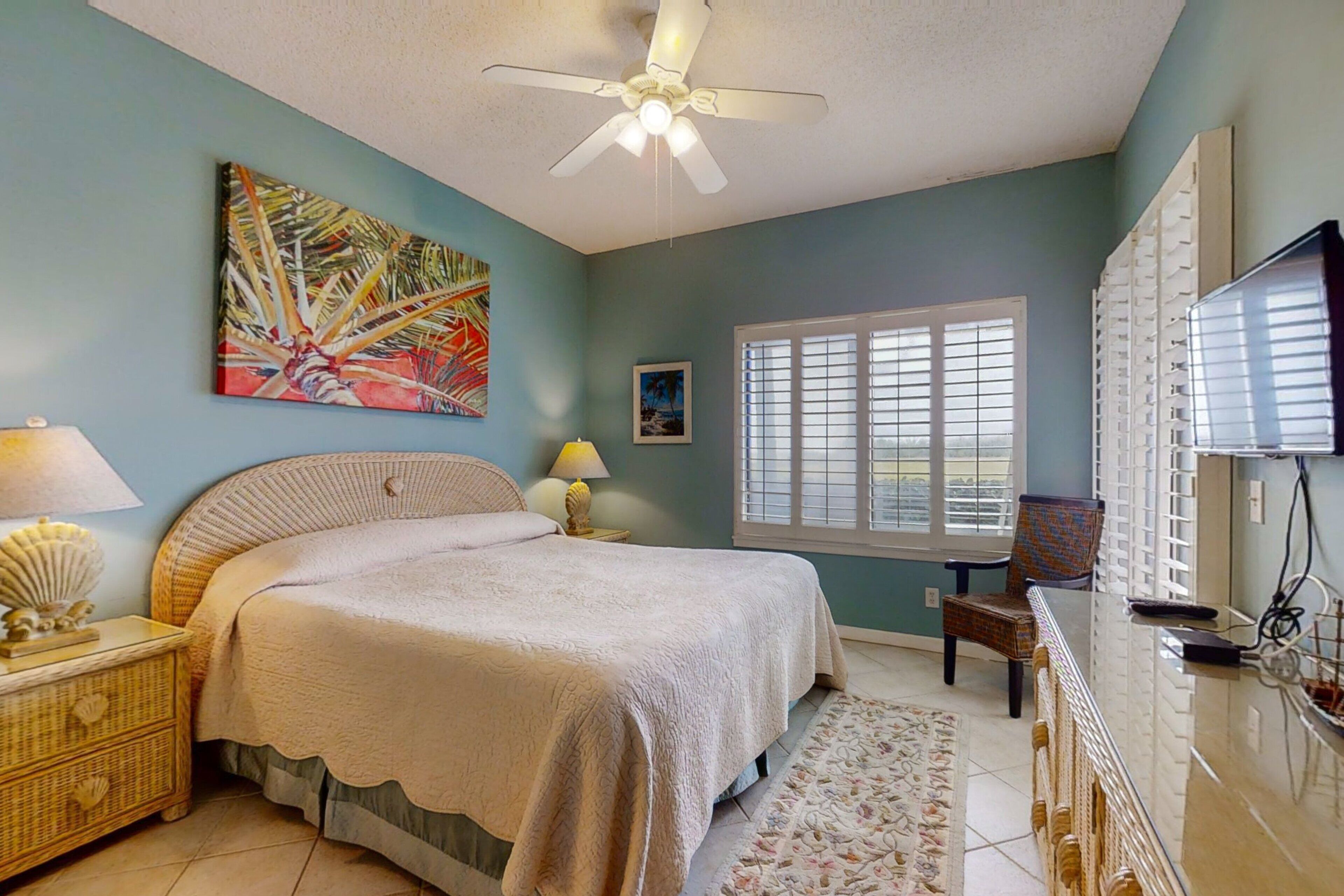 Une chambre à coucher comprenant un lit, un ventilateur de plafond, une fenêtre avec des stores, un téléviseur et un tableau décoratif.