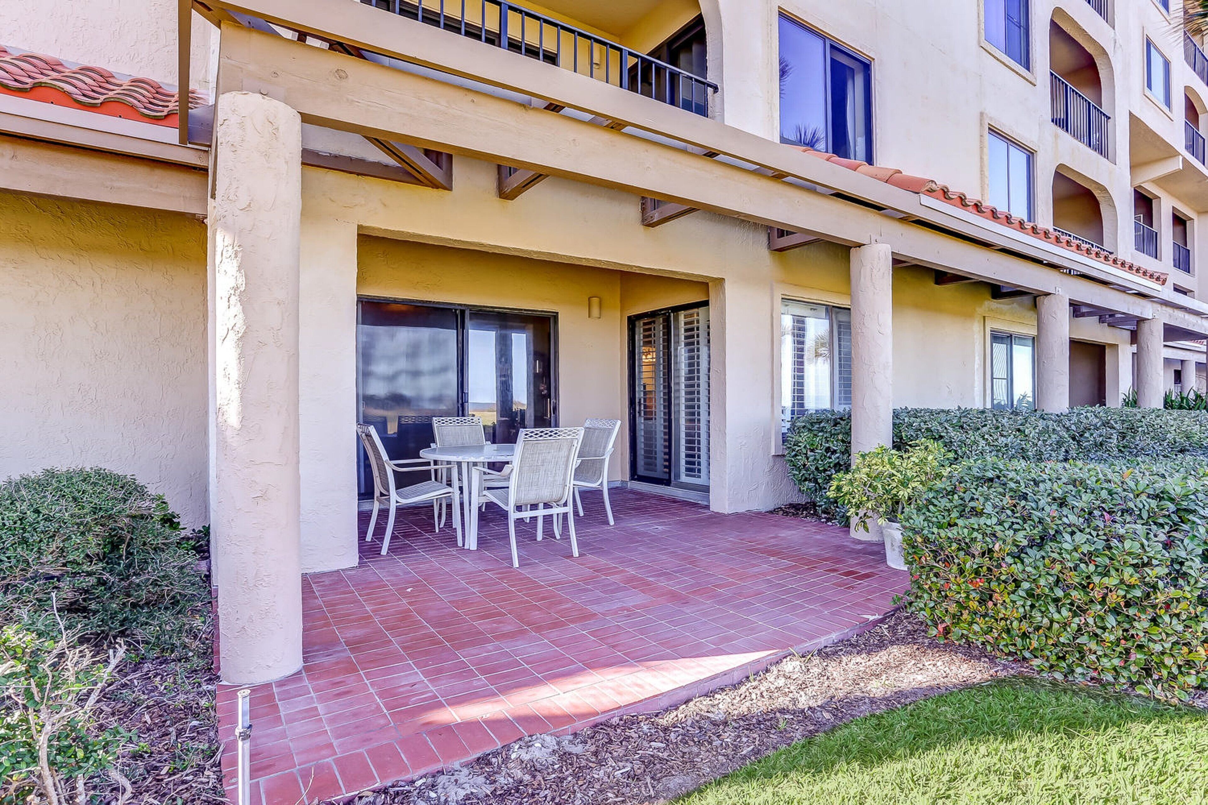 Un hotel con un patio cubierto que tiene una mesa y sillas, rodeado de vegetación y un sendero de baldosas.