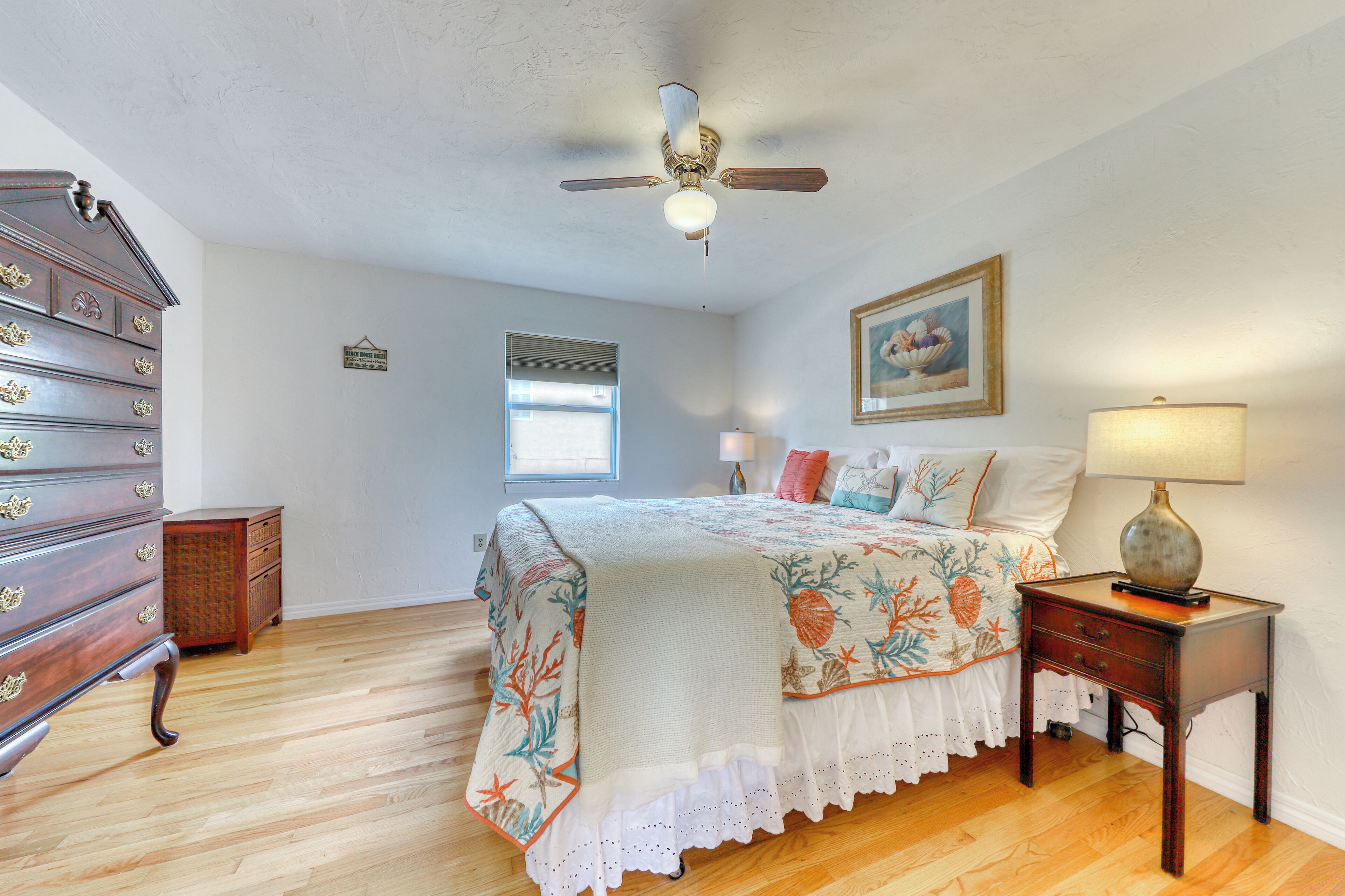 A bedroom with a wooden dresser, a bed with a floral bedspread, a ceiling fan, a nightstand with a lamp, and a window with blinds.