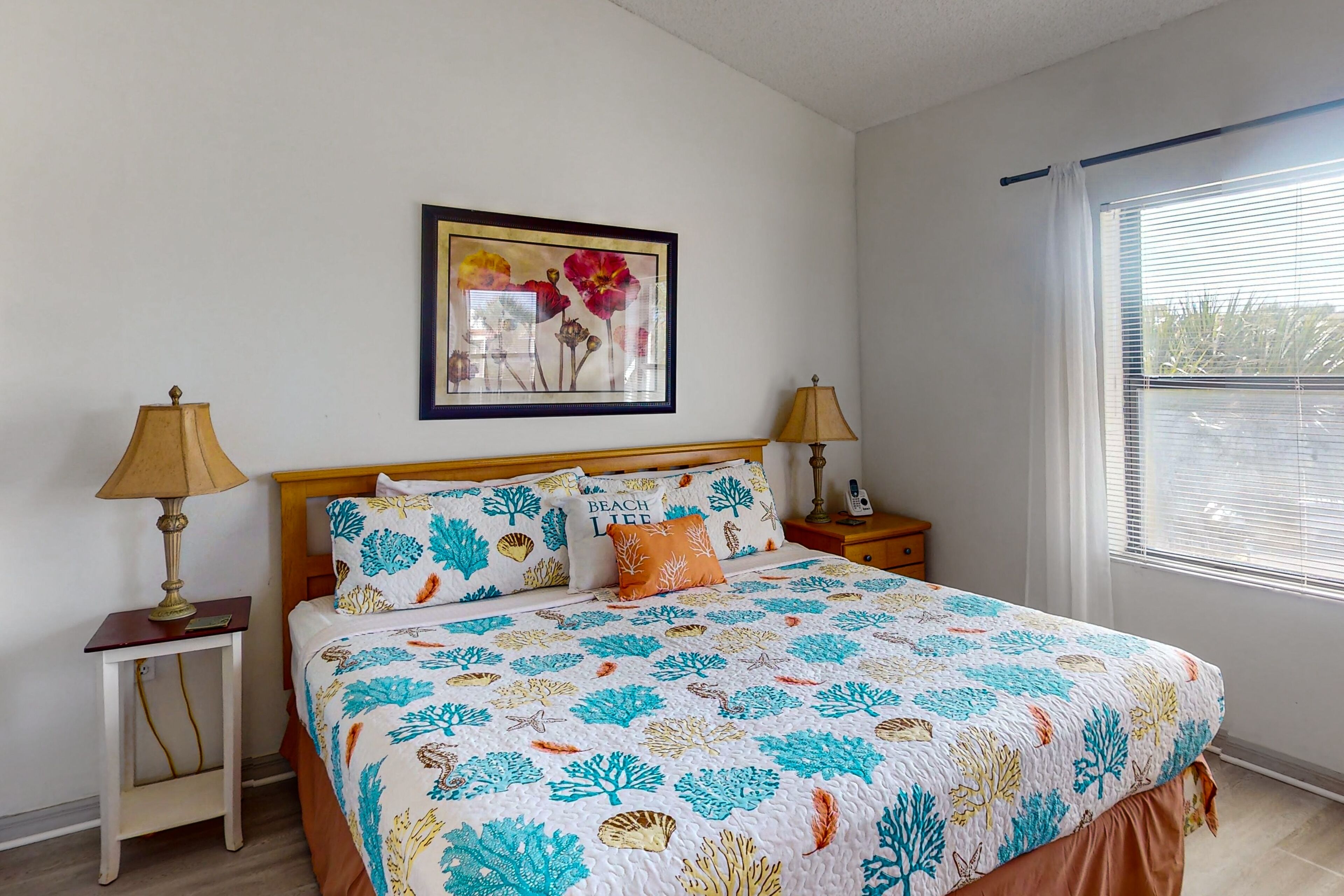 A bedroom with a bed, two bedside tables, a nightstand, a framed picture on the wall, and a window with blinds.