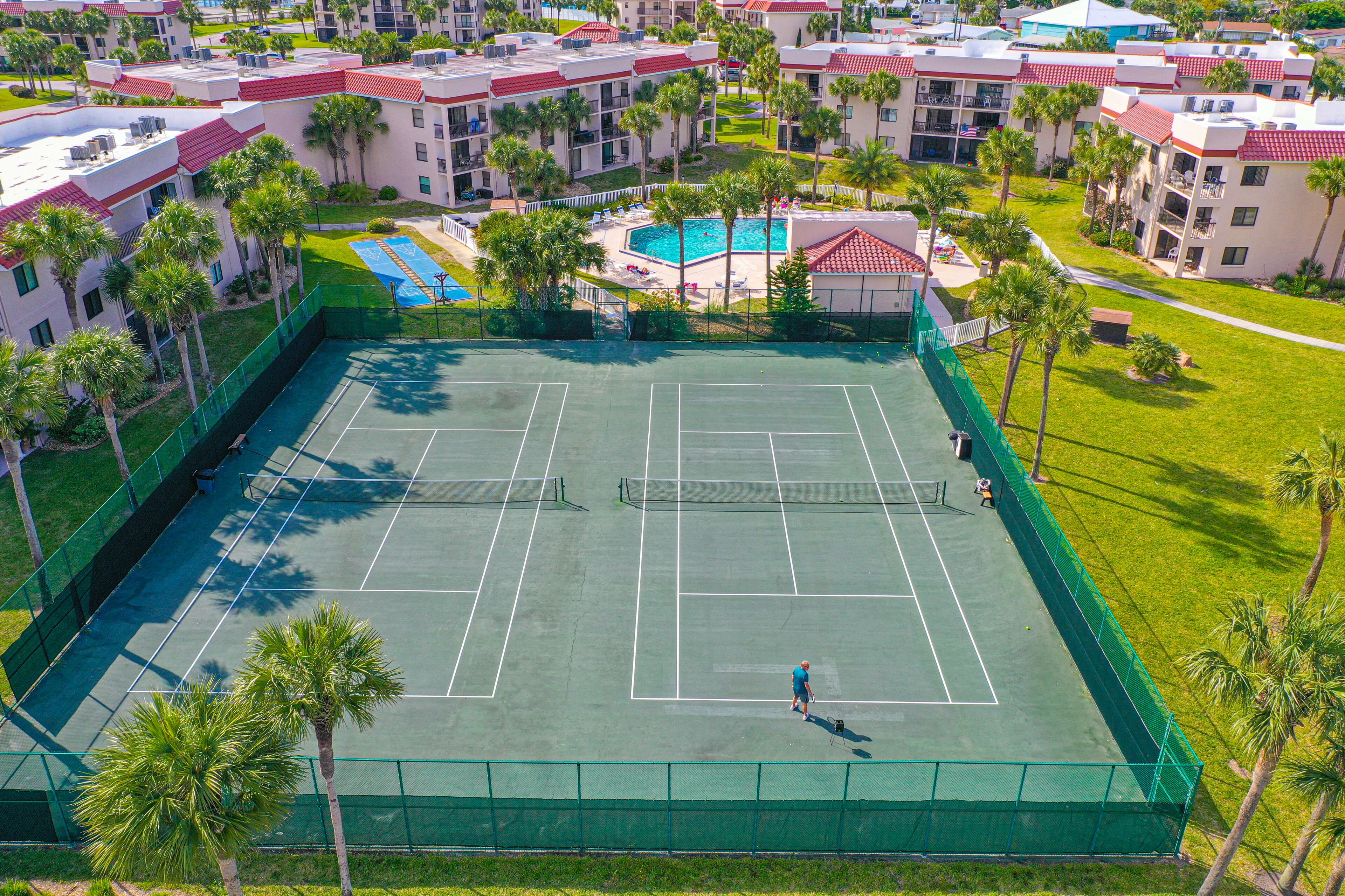 An aerial view of a tennis court surrounded by palm trees and a pool area, with a residential building in the background.