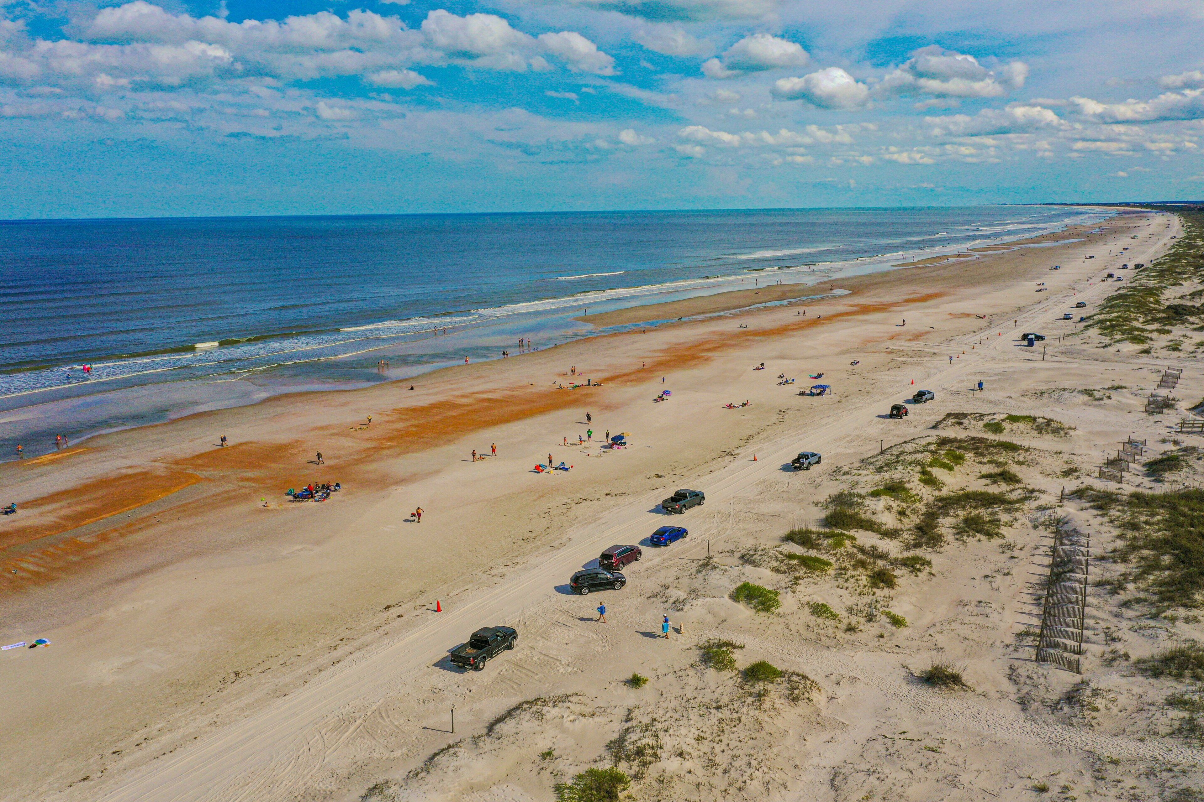 Een strand met een lange zandvlakte, enkele geparkeerde auto's en wat mensen die wandelen.