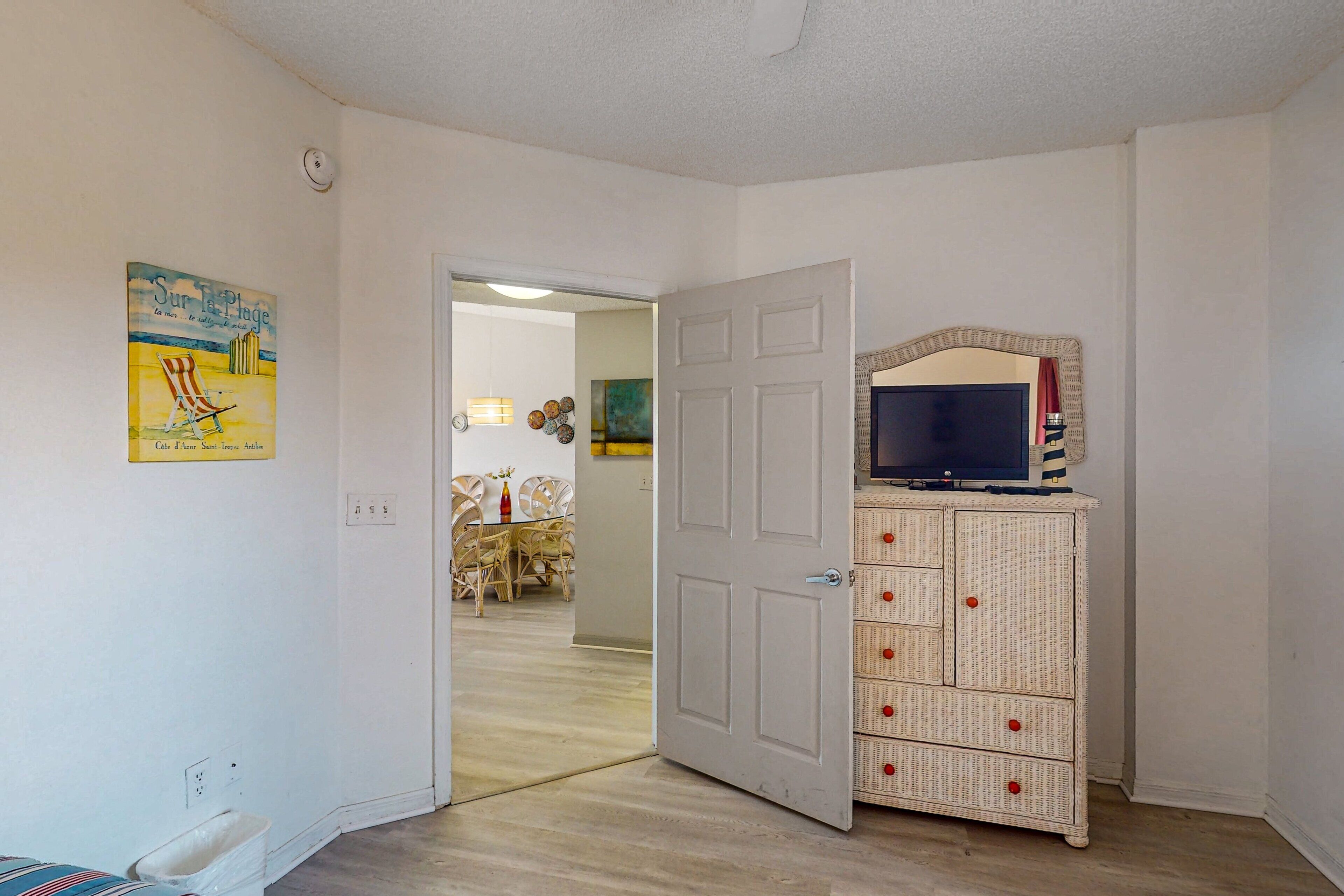 A room with a white door, a television on a cabinet, and a dining area in the background.