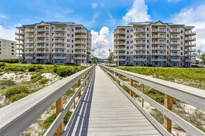 A wooden boardwalk leading to a beachfront apartment complex with multiple balconies and a clear sky. - Elegant, Oceanfront Condo With Balcony, Washer/dryer & On-site Pool (Fernandina Beach)