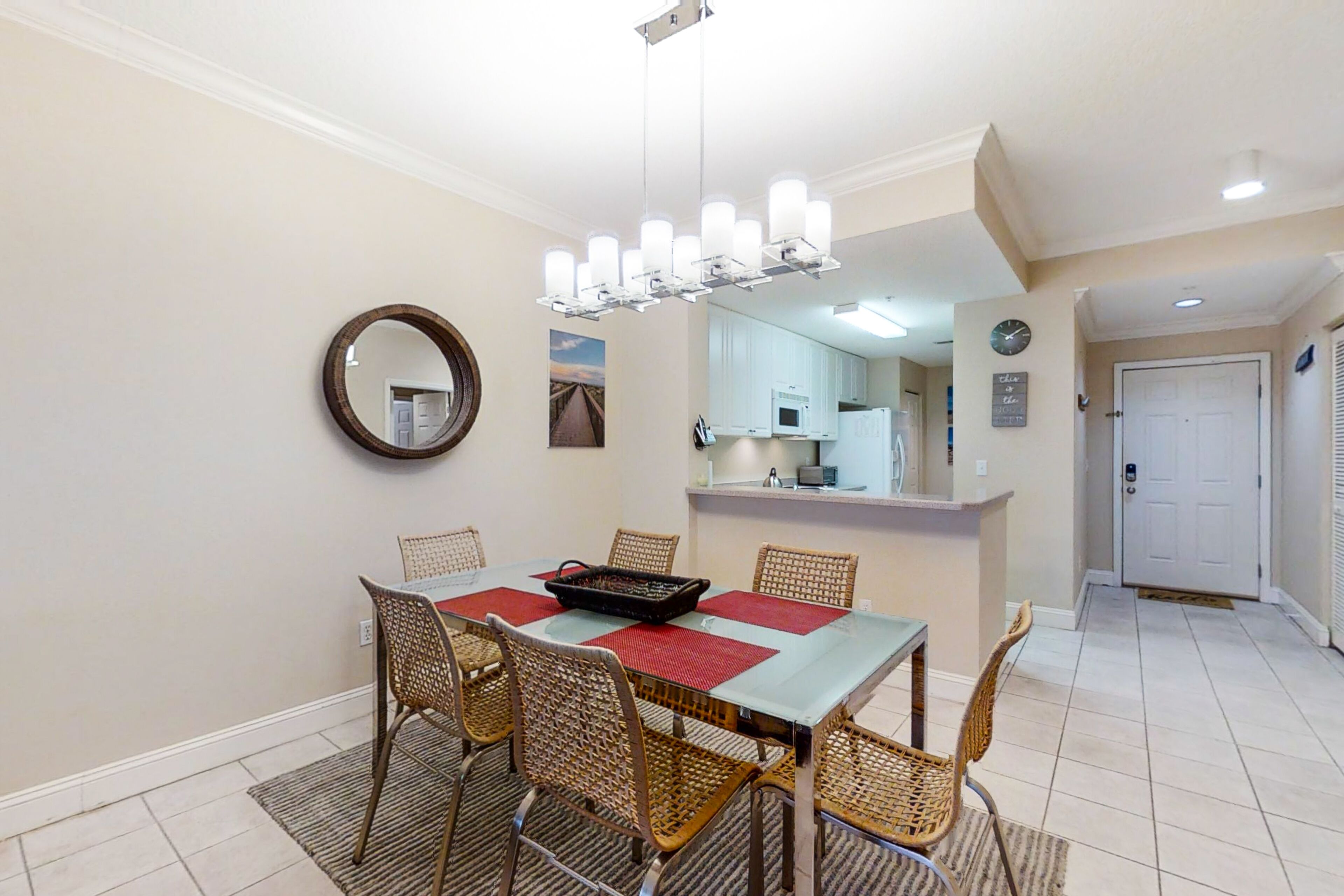 A dining area with a rectangular table, six wicker chairs, a chandelier, a mirror, and a kitchen in the background.
