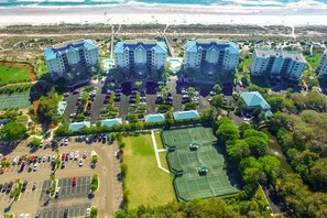 Um hotel moderno com edifícios de vários andares e telhados azuis, estacionamento, quadras de tênis e vista para a praia.