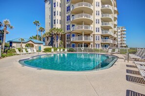 Uma piscina com espreguiçadeiras, um edifício com várias varandas e palmeiras.