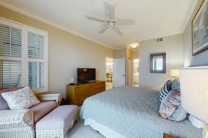 Um quarto com uma cama, televisão sobre uma cômoda, ventilador de teto e uma cadeira de vime.