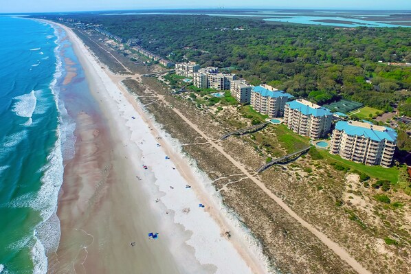 Vista aérea de uma área costeira com praia, um caminho para pedestres e vários edifícios altos com telhados azuis.
