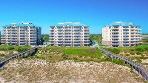 Tre edifici a più piani con balconi, una spiaggia sabbiosa in primo piano e una passerella in legno che li collega.