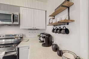 Una cocina con armarios blancos, un microondas de acero inoxidable, un hervidor, una cafetera y ollas colgantes.