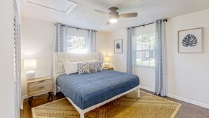 Une chambre à coucher comprenant un lit, des tables de chevet, un ventilateur de plafond et deux fenêtres avec des rideaux.