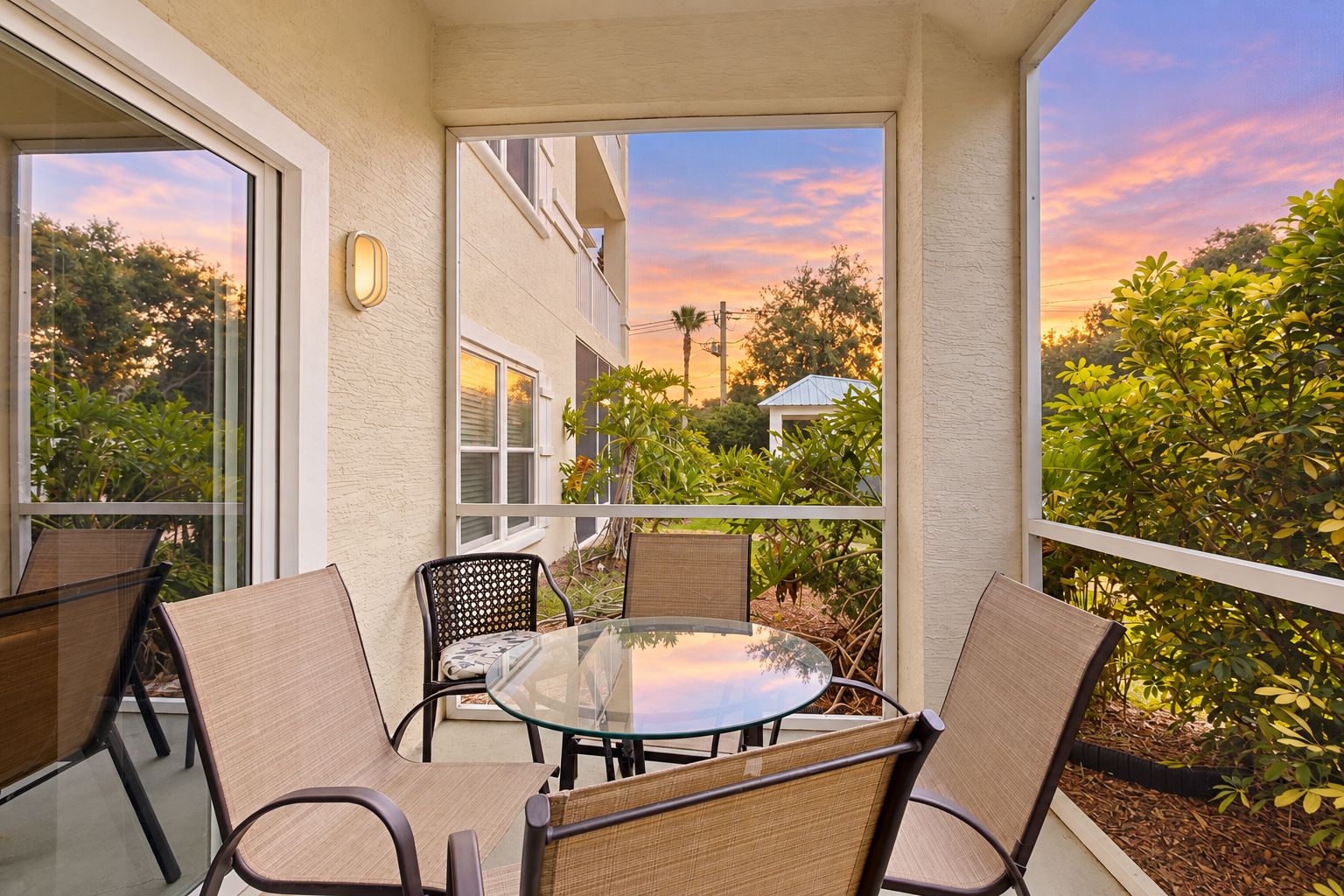 A glass-enclosed balcony with a round table and four chairs, overlooking a residential area with trees and a clear sky.