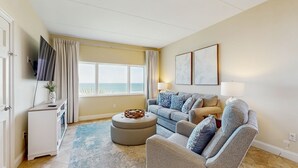 A living room with a grey sofa, a white cabinet, a round ottoman, and a large window with curtains. - Dual Suites & Ocean Views at Sandcastles Villas Pool, 2 Balconies & Golf (Fernandina Beach)