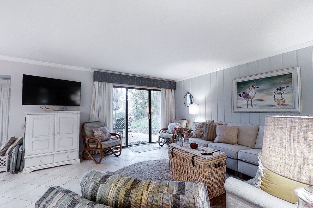 A spacious living room with a large flat-screen TV, a sofa set, a wicker coffee table, and a painting on the wall.