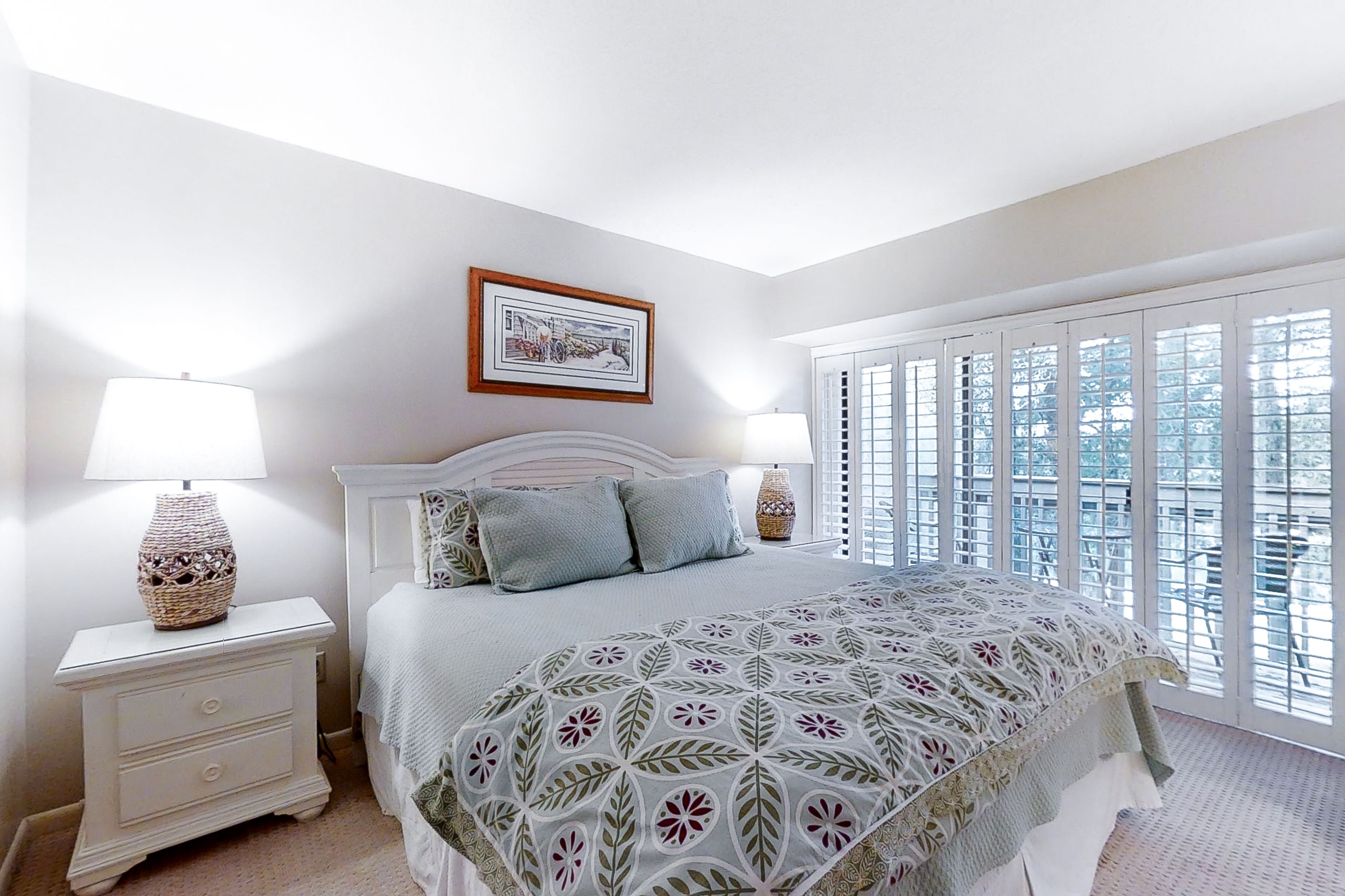 A bedroom with a bed, nightstands, a lamp, a framed picture, and a window with blinds.