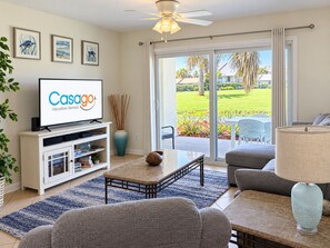 A living room with a flat-screen TV displaying 'Casago Vacation Rentals,' a ceiling fan, a sliding glass door leading to a patio, and a blue rug. - Garden-level Condo With Patio, 2 Pools, Gym, Tennis & Sauna - Steps to the Beach (St. Augustine)