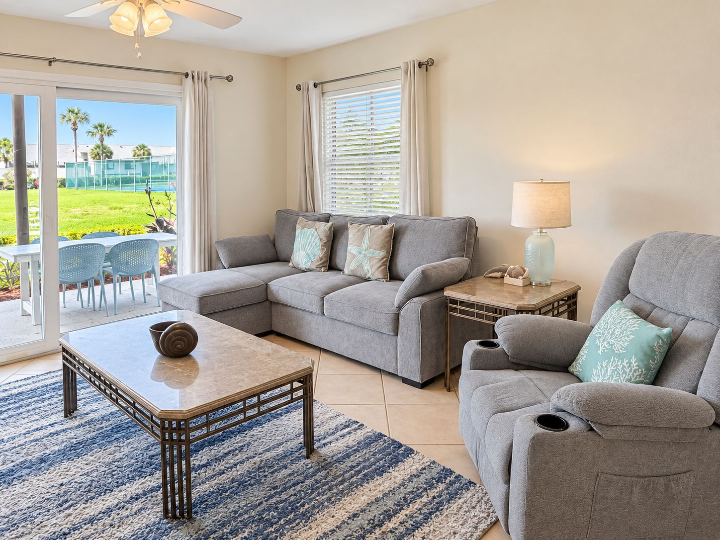 A living room with a grey sectional sofa, a small table, and a chair. There is a window with blinds, a lamp, and a view of a green lawn.