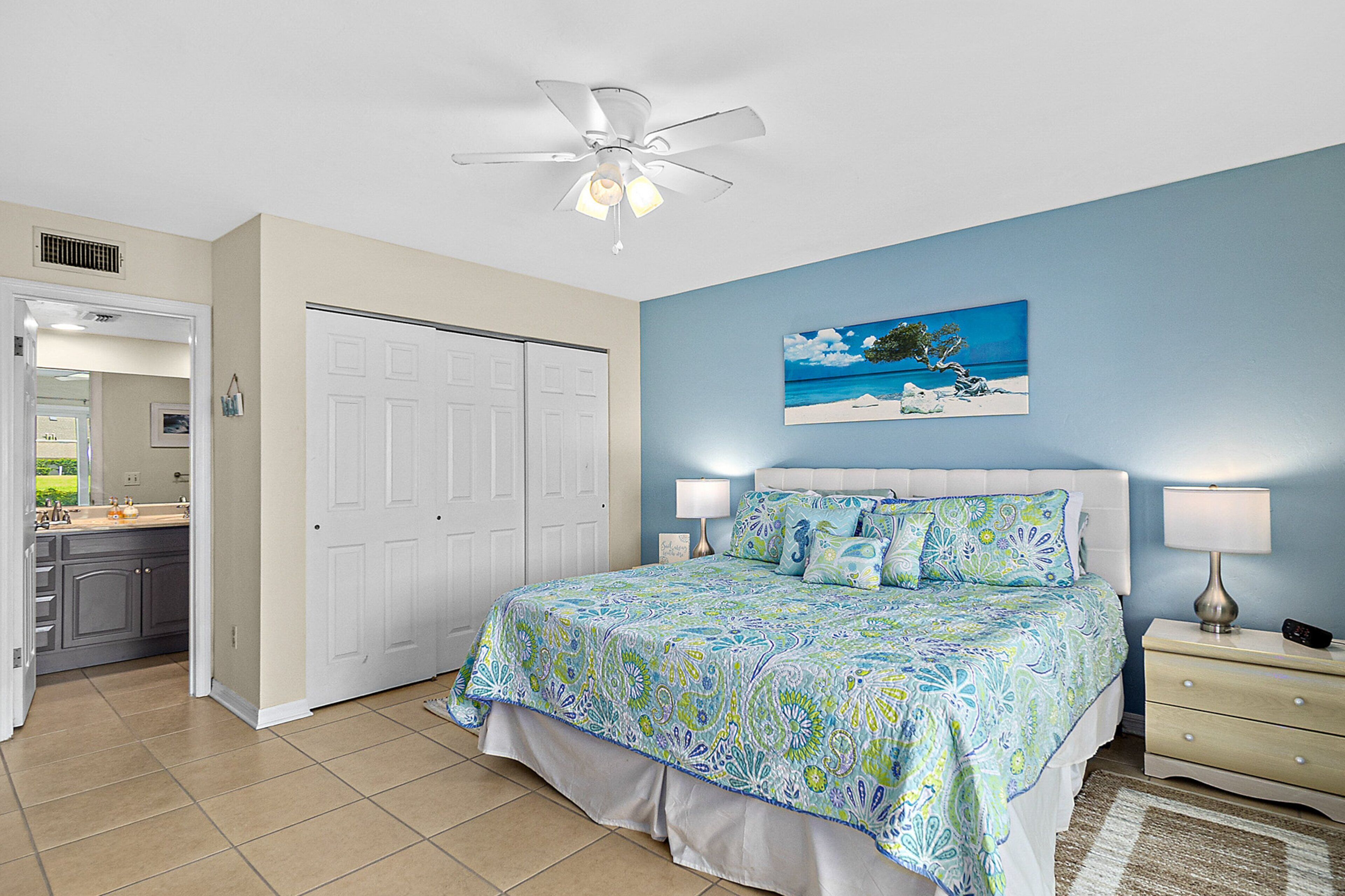 A bedroom with a bed, nightstands, a ceiling fan, a painting, and a bathroom with a sink and mirror.