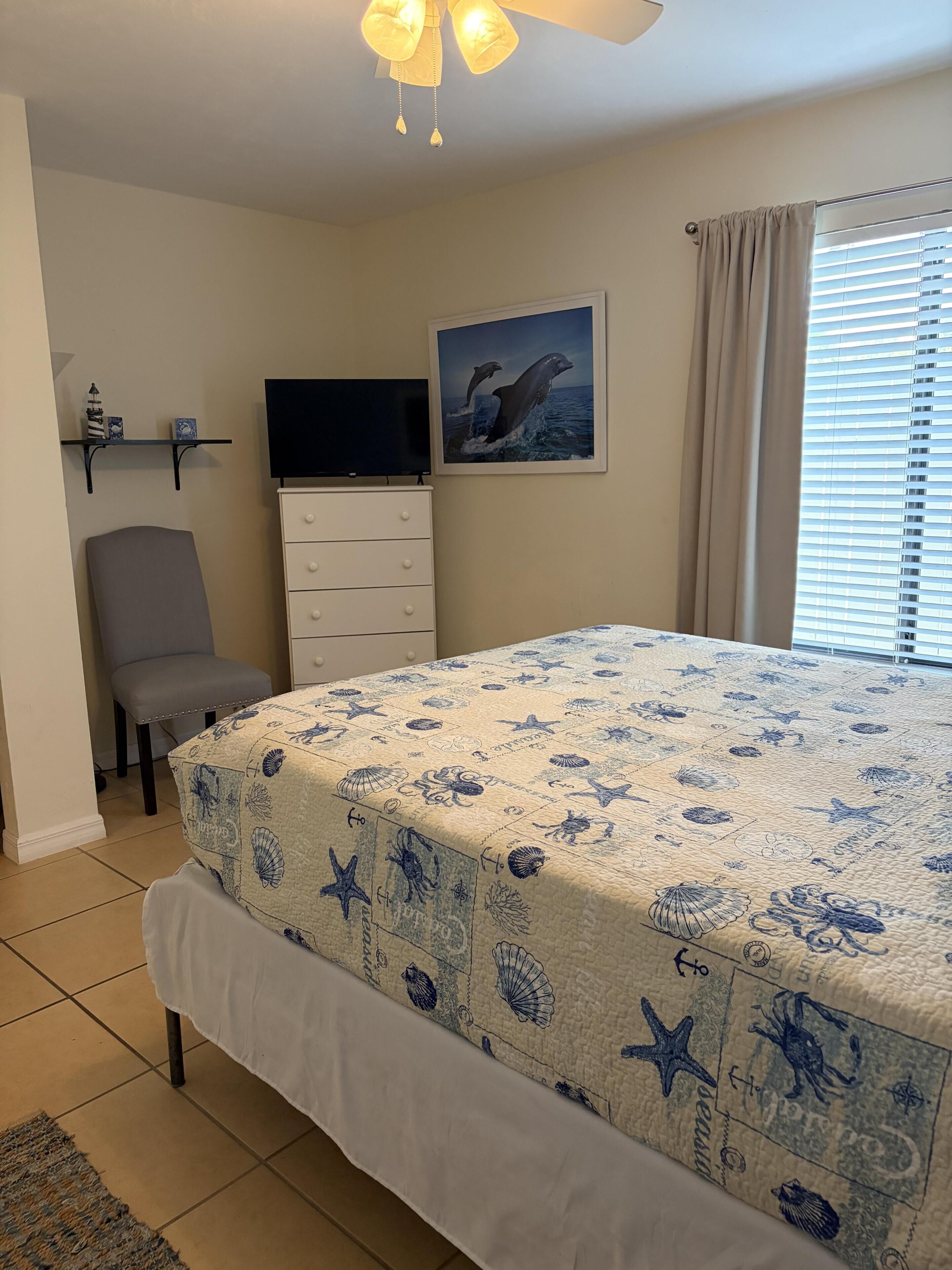 A bedroom with a bed, a chair, a TV, a dresser, a painting, and a window with blinds.