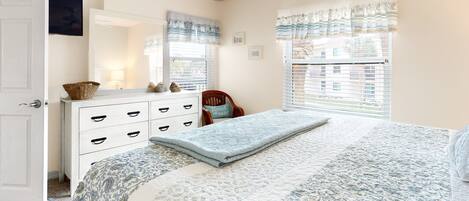 A bedroom with a bed, dresser, chair, and windows with curtains.