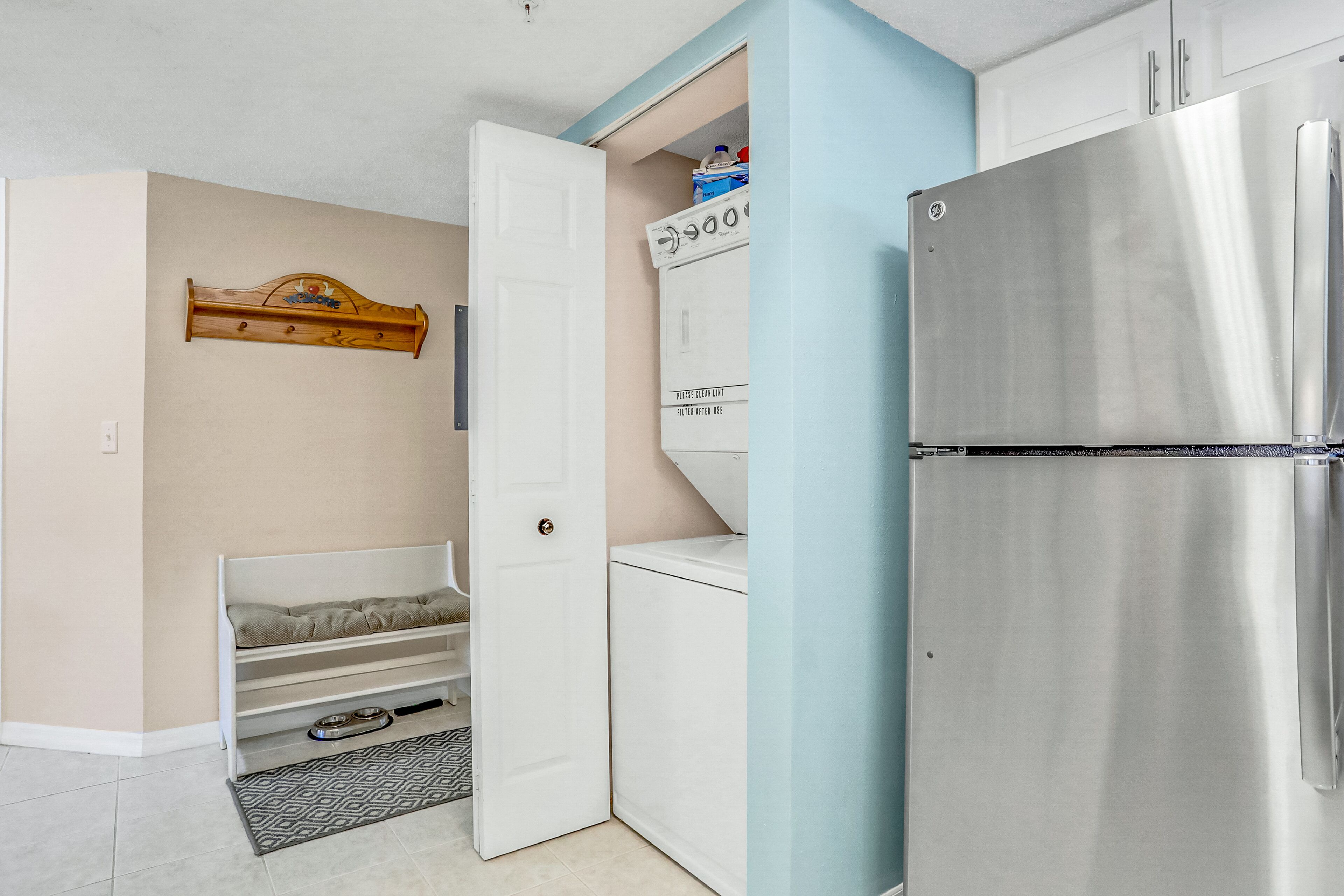 A compact kitchen with a built-in stainless steel refrigerator, a washing machine, and a small wooden shelf above a white door.