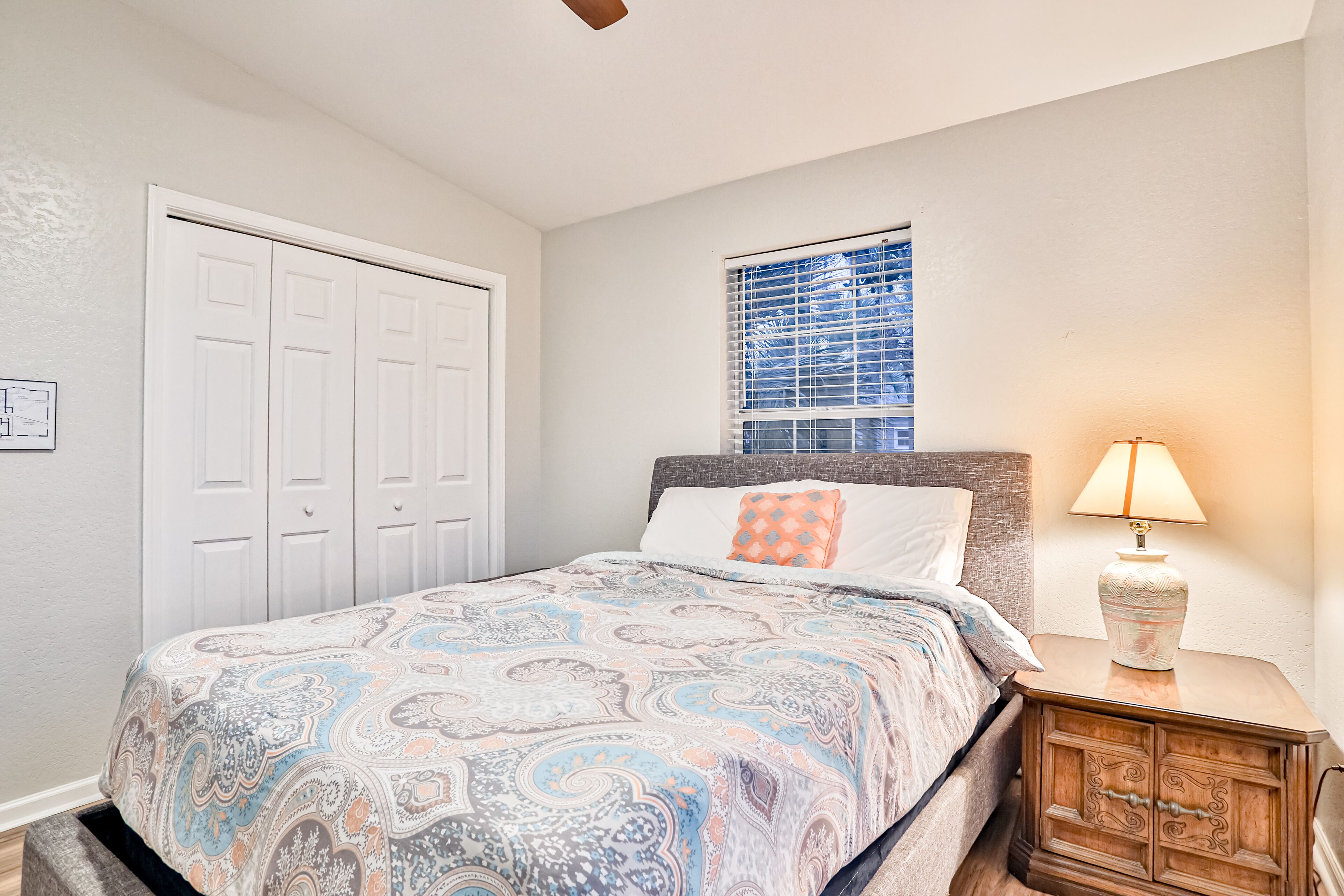 A bedroom with a bed, a nightstand with a lamp, a window with blinds, and a white door.