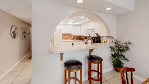 Eine Küche mit weißen Schränken, einer zentralen Kochinsel mit Hockern, einem Deckenventilator und einem Blumentopf.