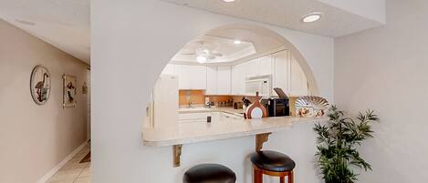 A kitchen with white cabinets, a central island with stools, a ceiling fan, and a potted plant.