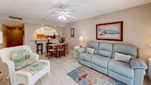 Ein Wohnzimmer mit einem hellblauen Sofa, einem weißen Sessel, einem Essbereich mit Tisch und Stühlen sowie einem Deckenventilator.