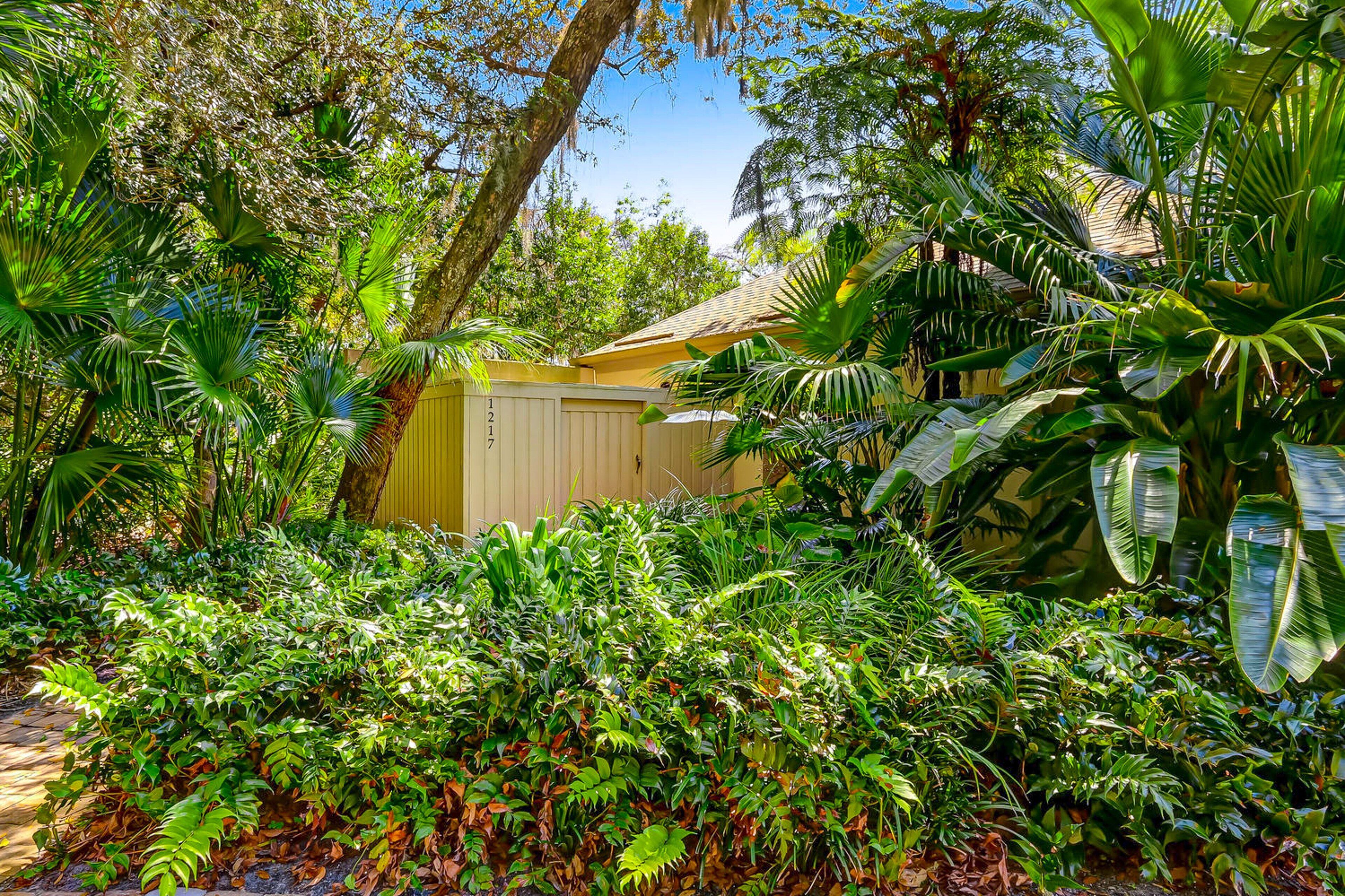 A lush garden with dense foliage, a yellow fence, and a building with a tiled roof.