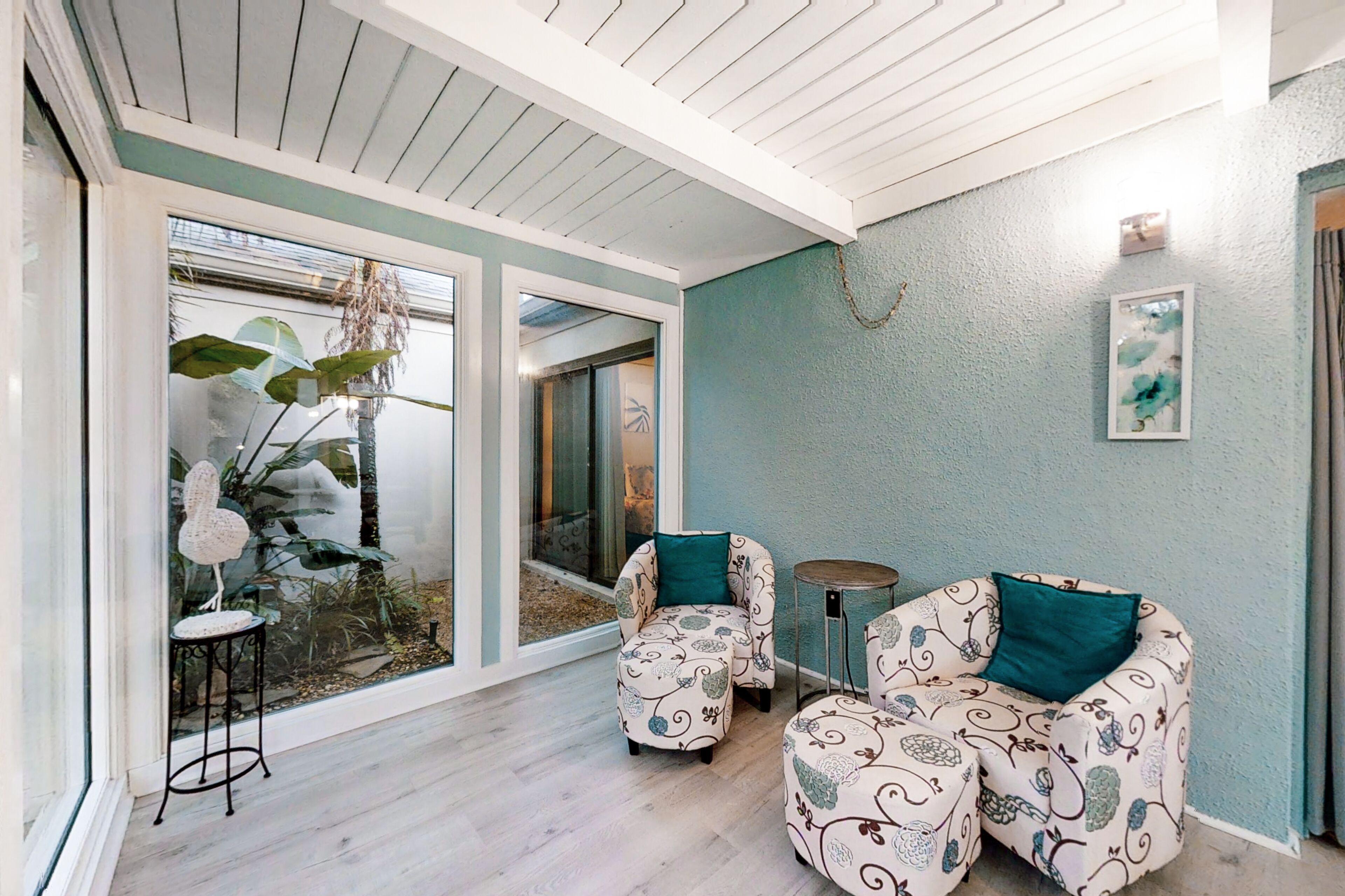Una sala de estar moderna con un amplio ventanal corredera que da a un jardín, un conjunto de sofá con cojines estampados y una pequeña mesa redonda.