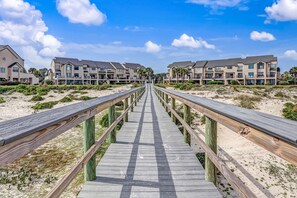 Une promenade en bois mène à une propriété située en bord de mer, avec des immeubles à plusieurs étages, dont certains sont dotés de balcons.