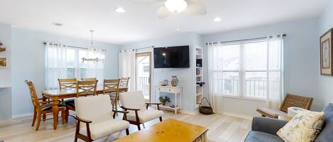 A spacious living room with a dining area, a ceiling fan, a flat-screen TV, and large windows.