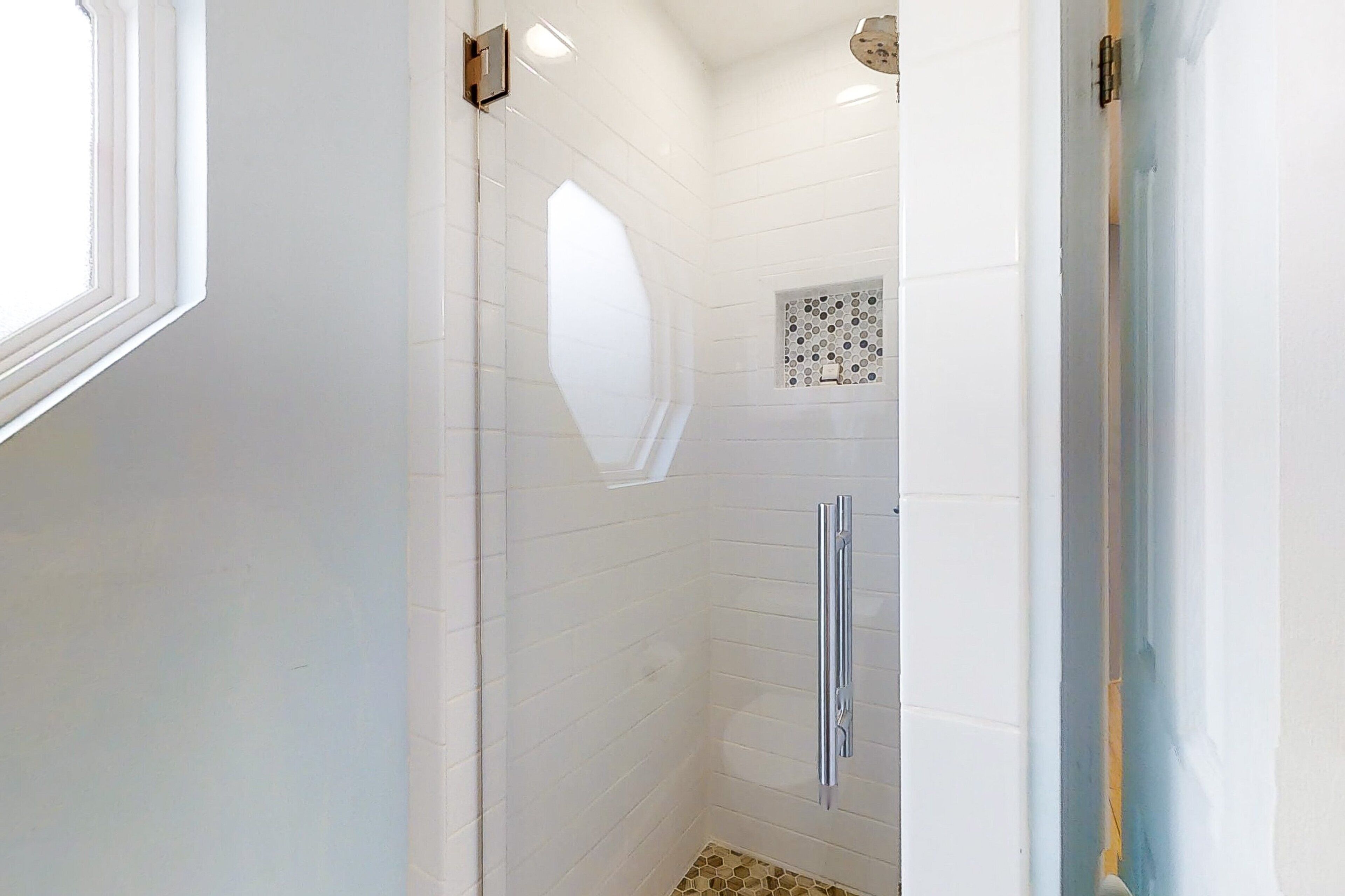 Un baño en esquina con paredes cubiertas de azulejos blancos, una ducha con puerta de vidrio y un ventanal pequeño.