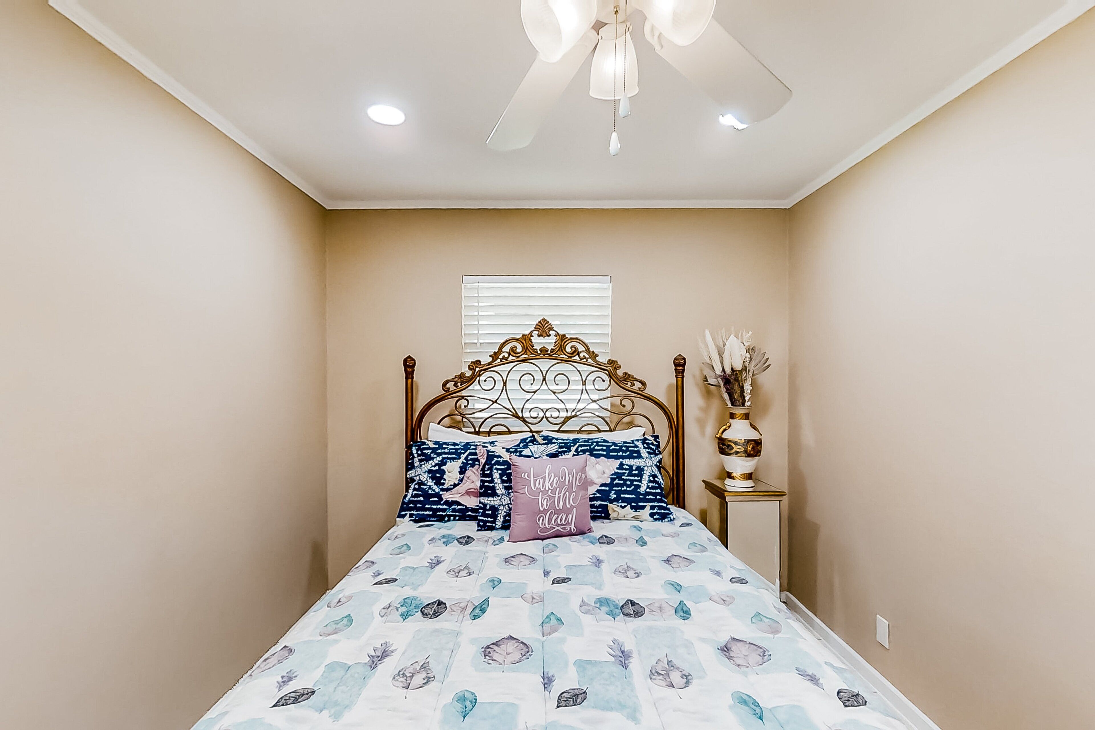 A bedroom with a bed, a ceiling fan, a window with blinds, and a bedside table with a vase.