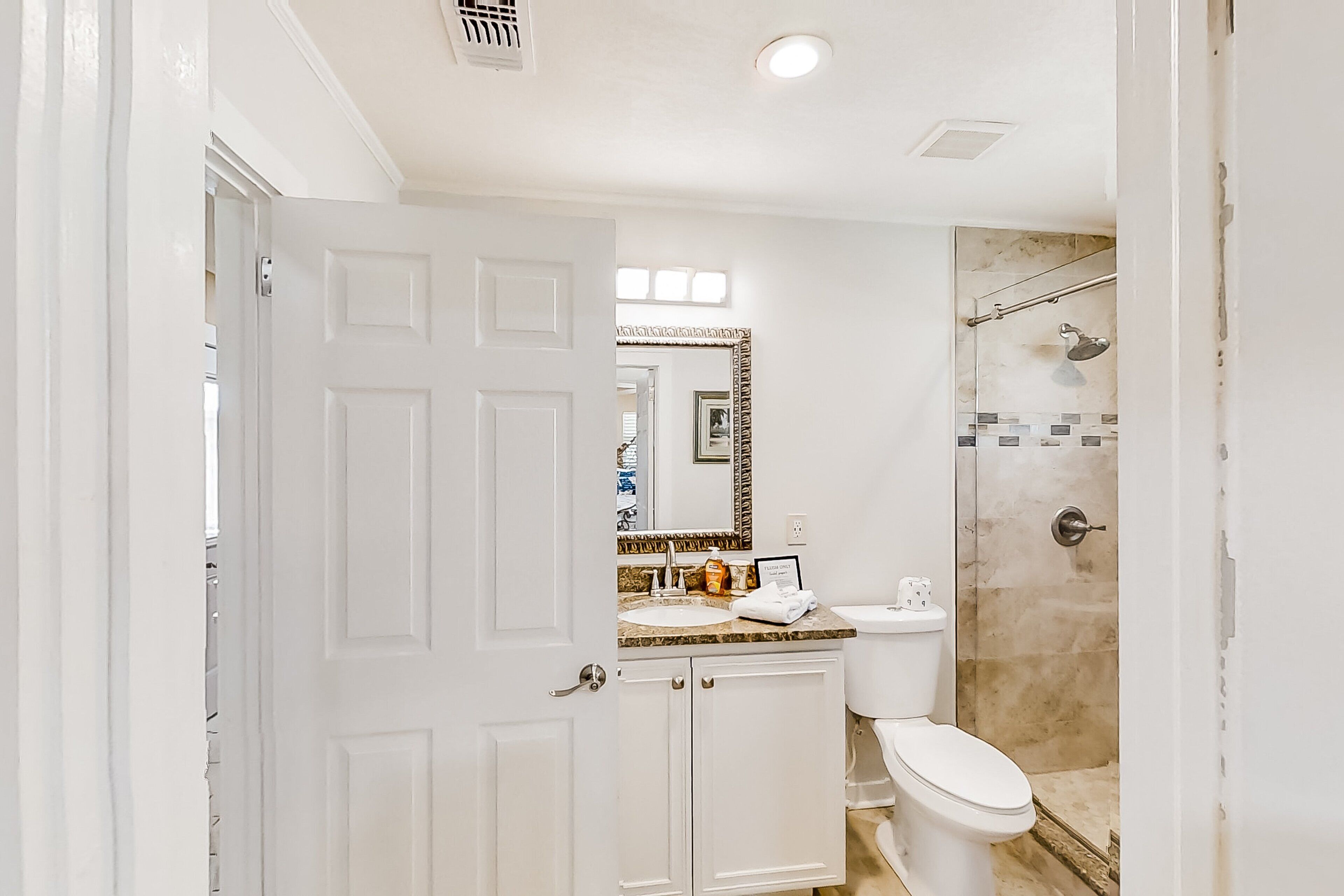 A bathroom with a white door, a sink with a mirror above it, a toilet, and a shower area.