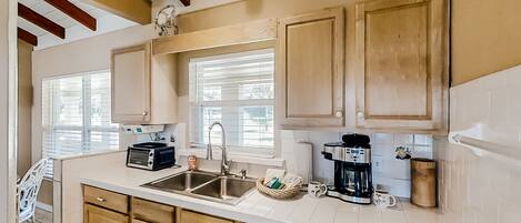 A kitchen with wooden cabinets, a double sink, a coffee maker, and a window with blinds.