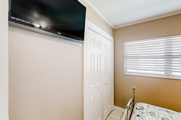 A bedroom with a TV mounted on the wall, a white wardrobe, a bed with a patterned cover, and a window with blinds. - Home Near the Beach & Golf With Inviting Patio, Fireplace, AC, & Washer/dryer (Fernandina Beach)