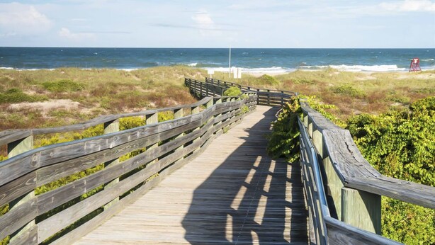 Ein Holzsteg führt zum Strand, in der Ferne ist ein roter Rettungsschwimmerstuhl zu sehen.