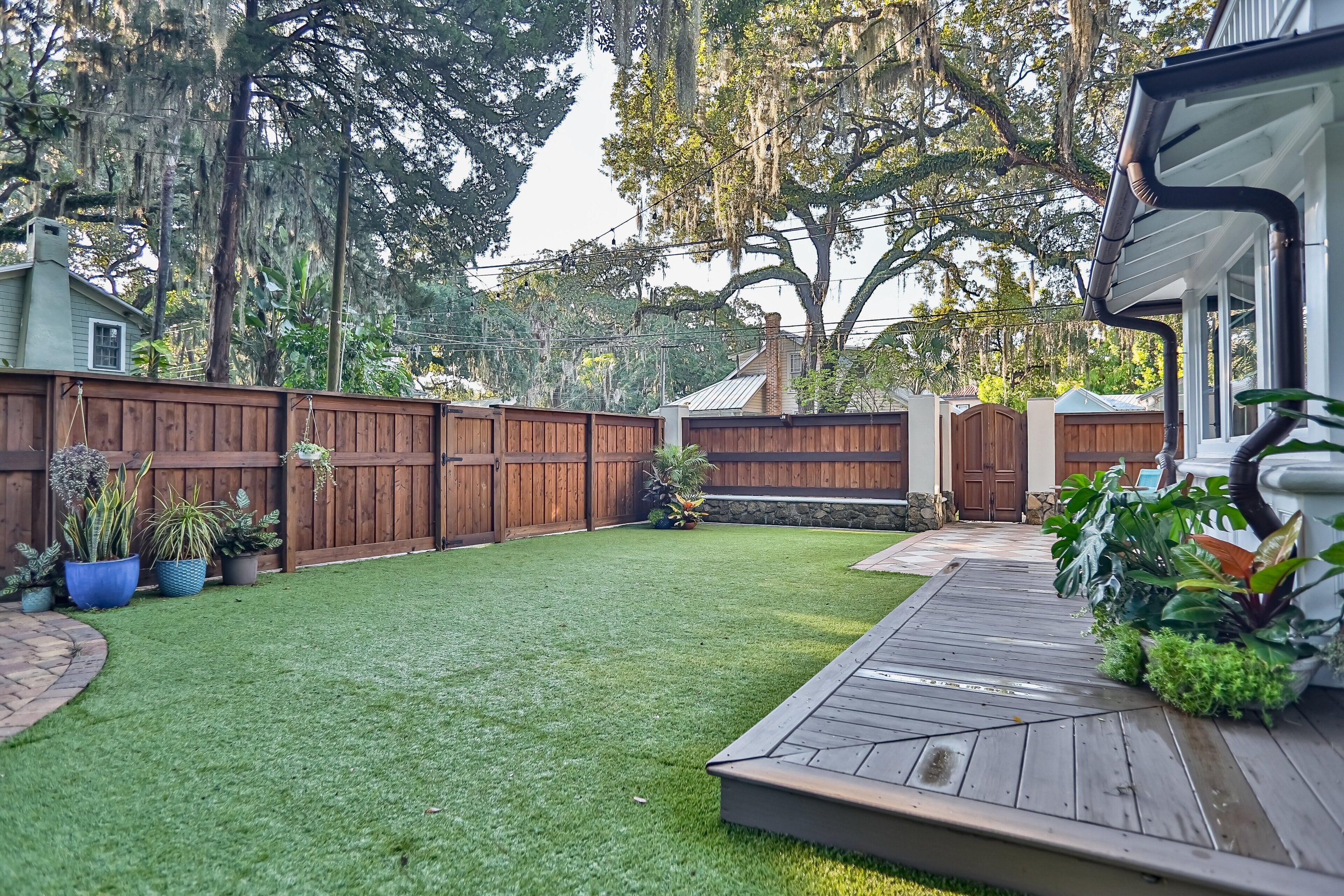 Un jardin avec une terrasse en bois, un espace clôturé et diverses plantes en pot.