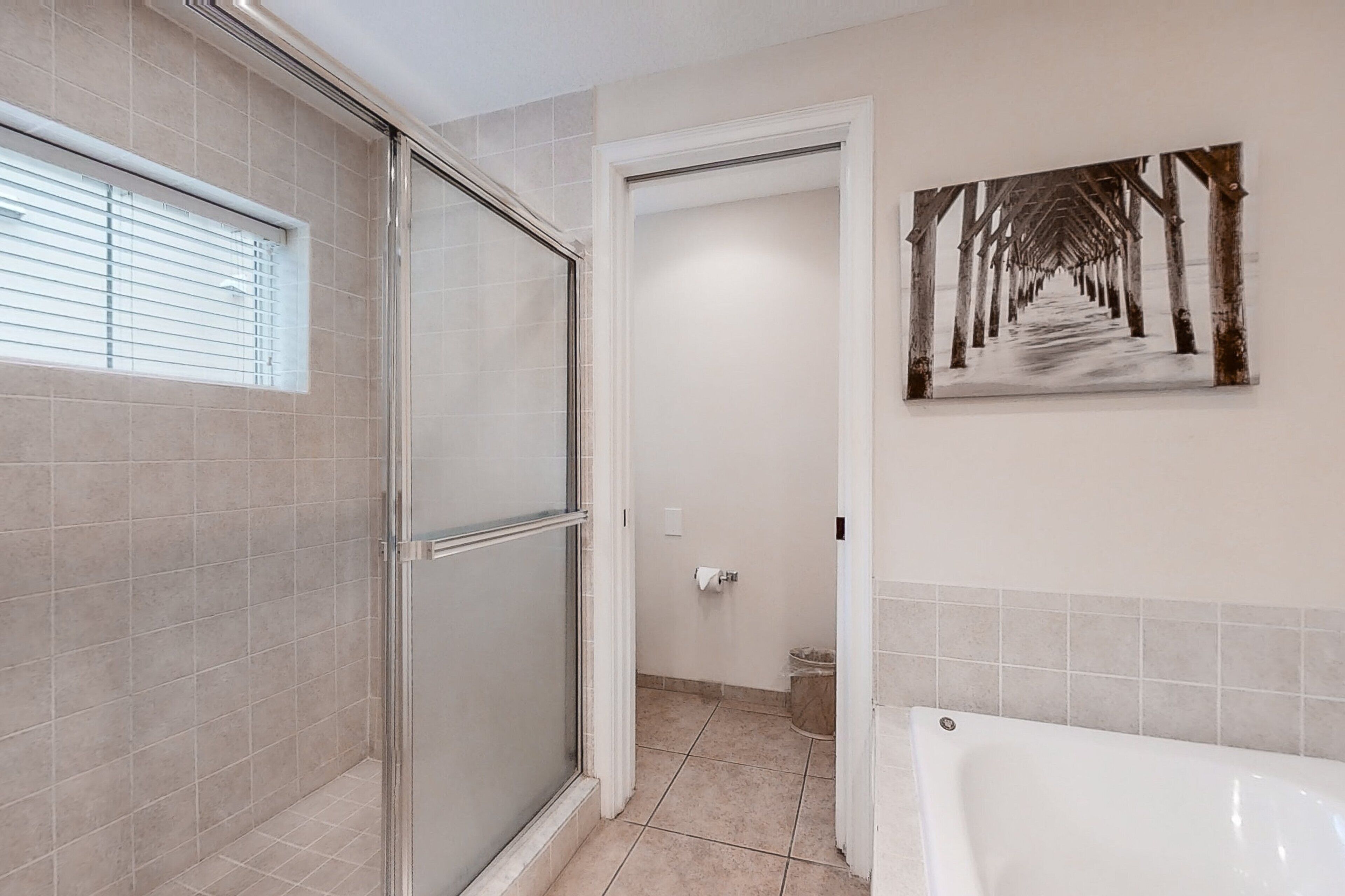 A bathroom with a glass shower enclosure, a white bathtub, and a painting of a pier on the wall.