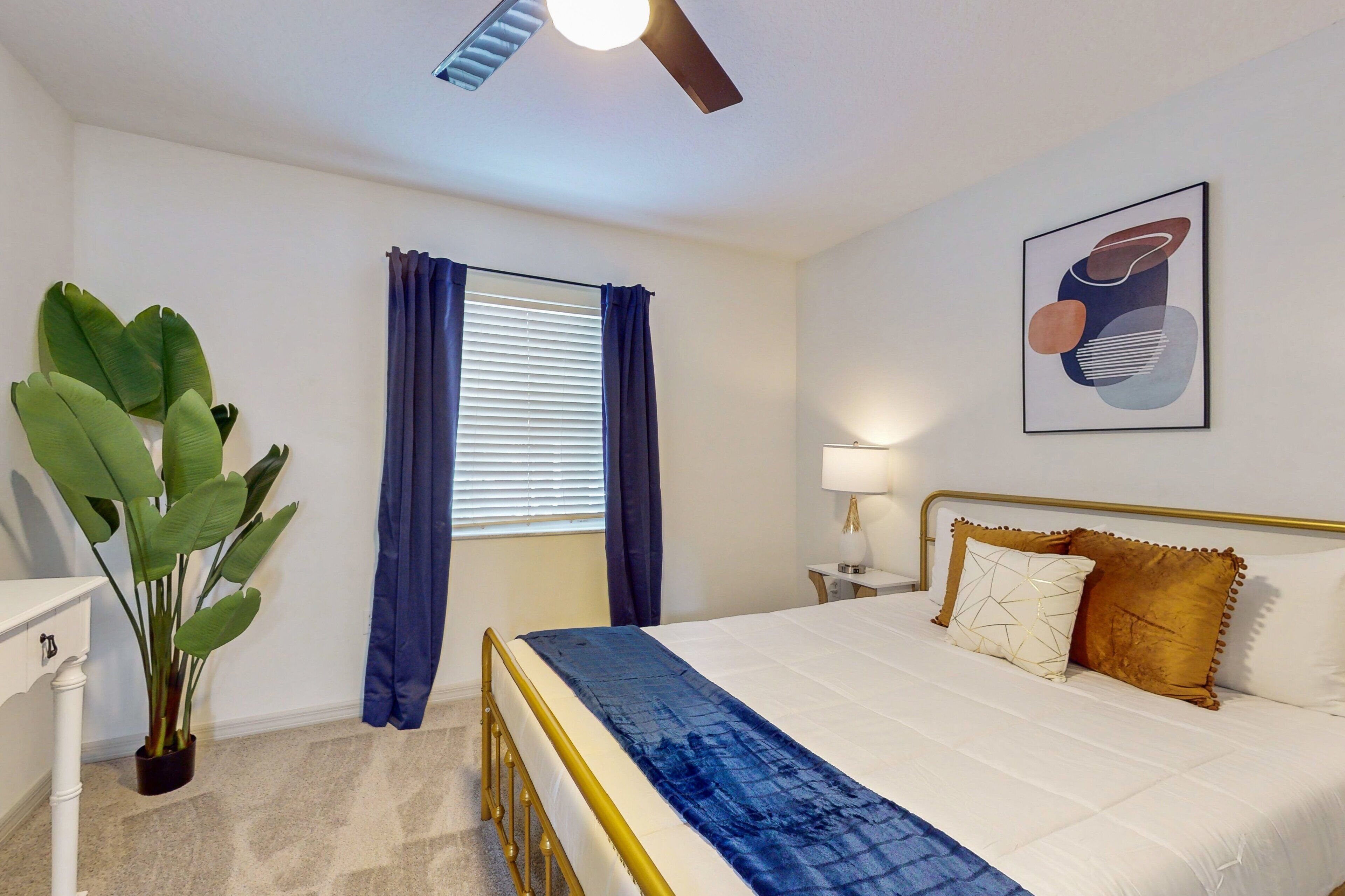 A bedroom with a bed, a nightstand, a lamp, a ceiling fan, and a large plant.